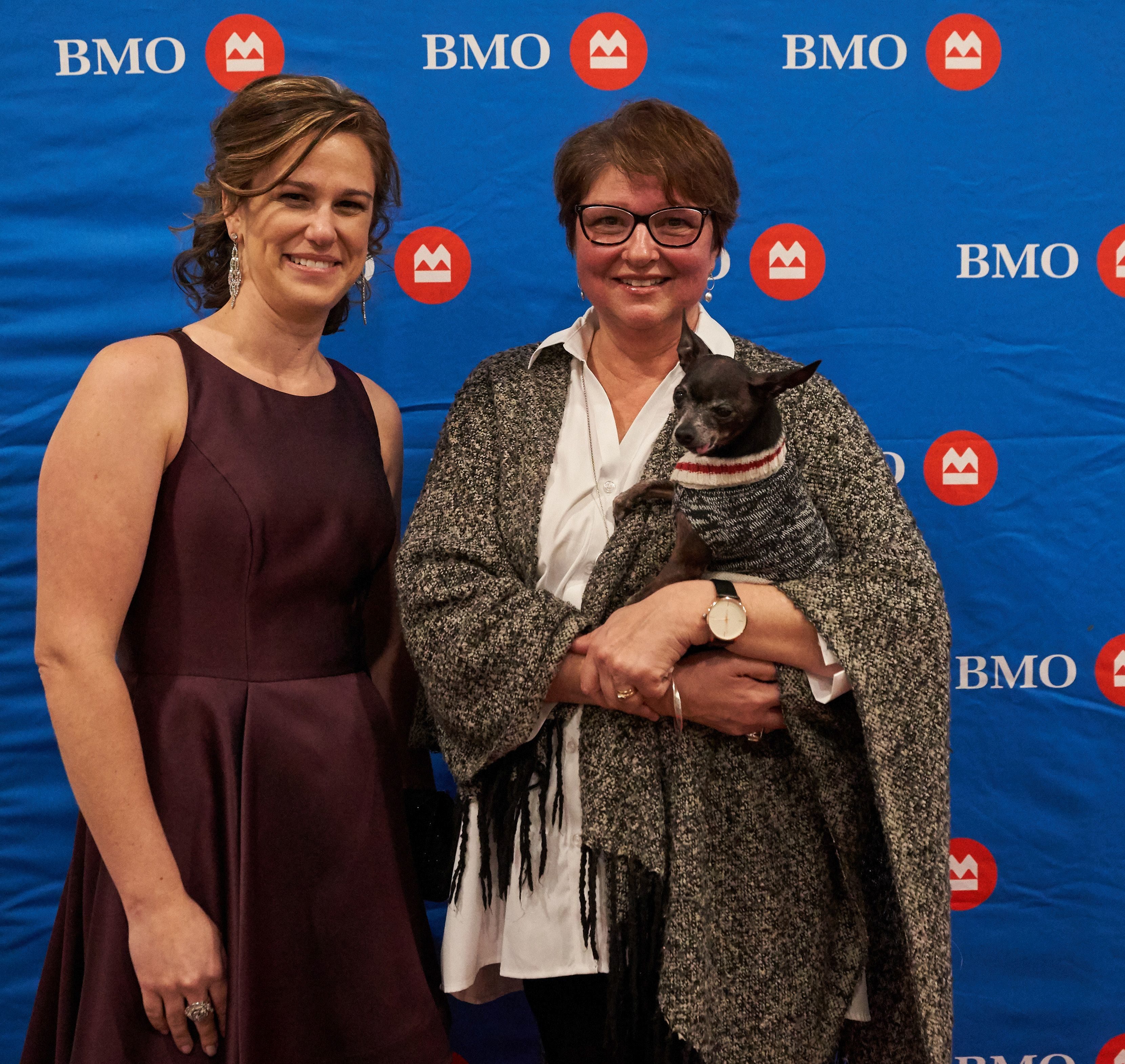Two women posing in front of a BMO backdrop at the gala, one holding a small black dog