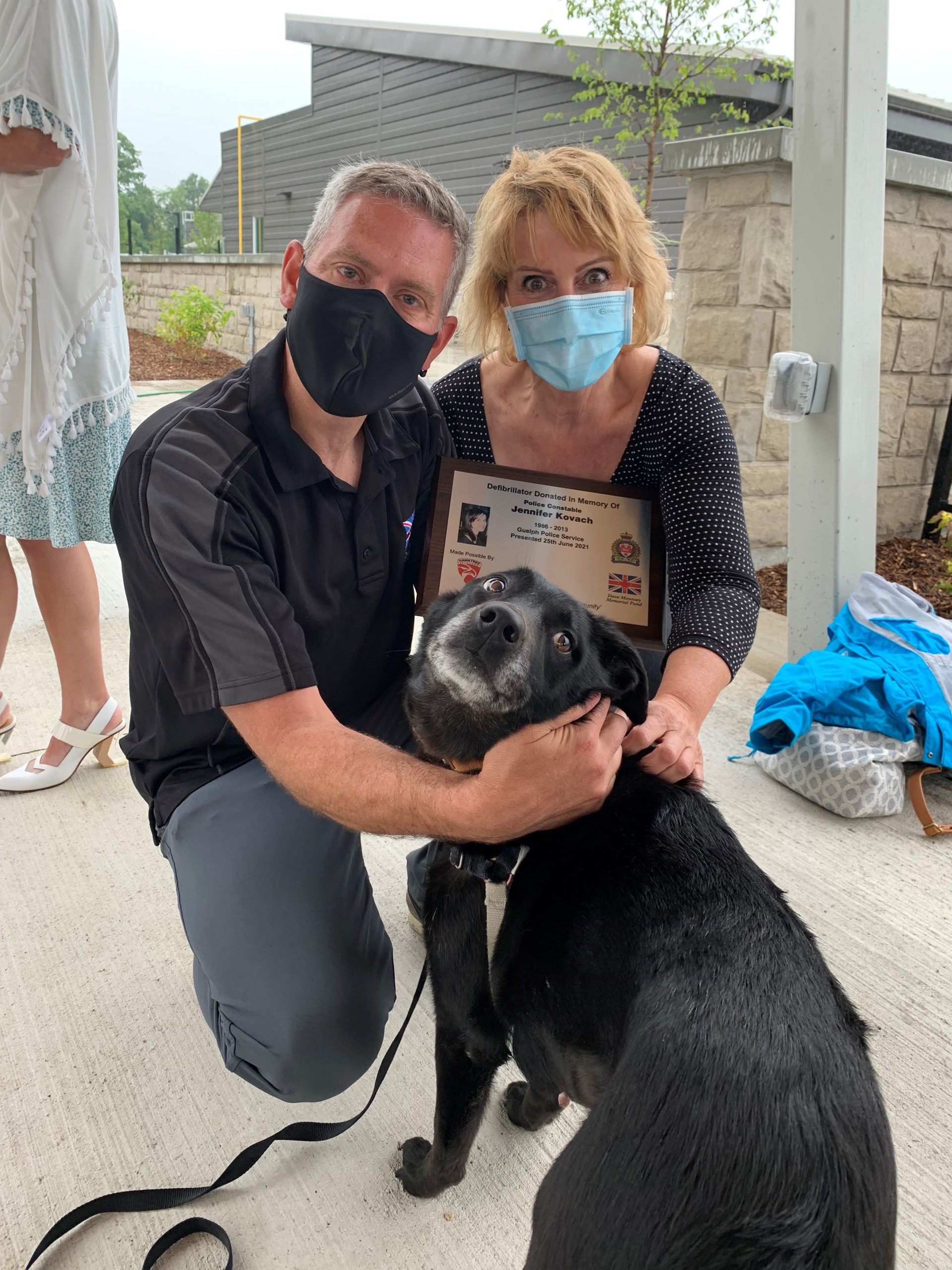 Patrick and Gloria, wearing face masks, kneeling with their black dog, Sylvie, and holding a plaque