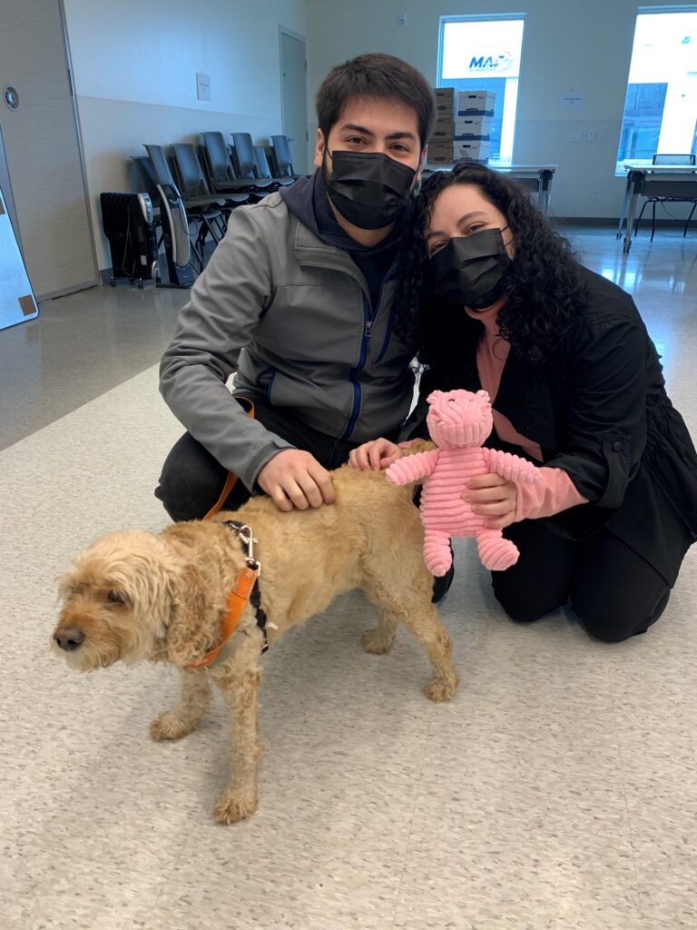 Betsy, a scruffy tan dog, with her new family, who are wearing face masks and holding a pink toy