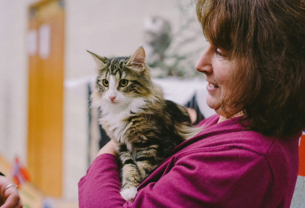 A woman in a magenta cardigan holds a long-haired tabby cat in her arms and smiles down at it.