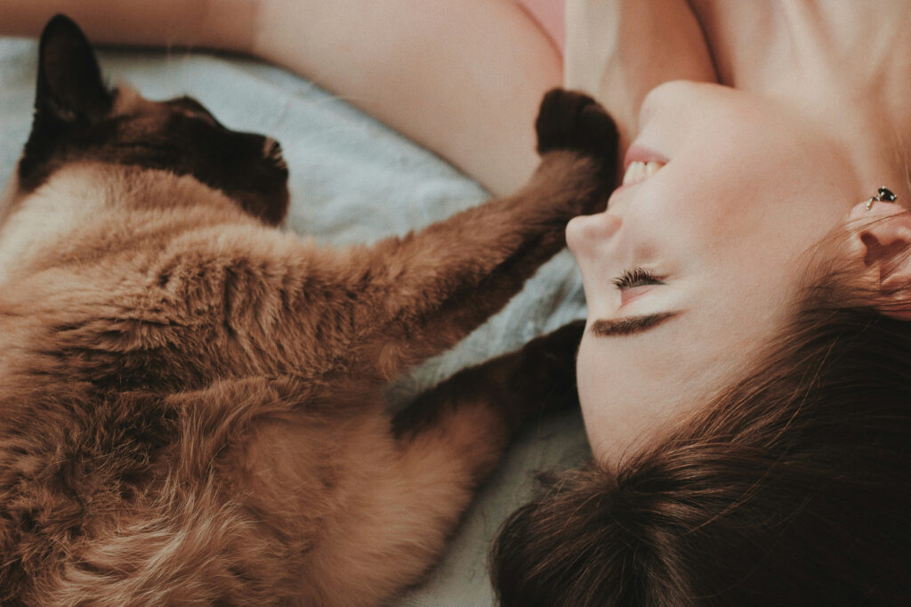 A woman lies on a bed laughing as a siamese cat stretches a paw toward her face.