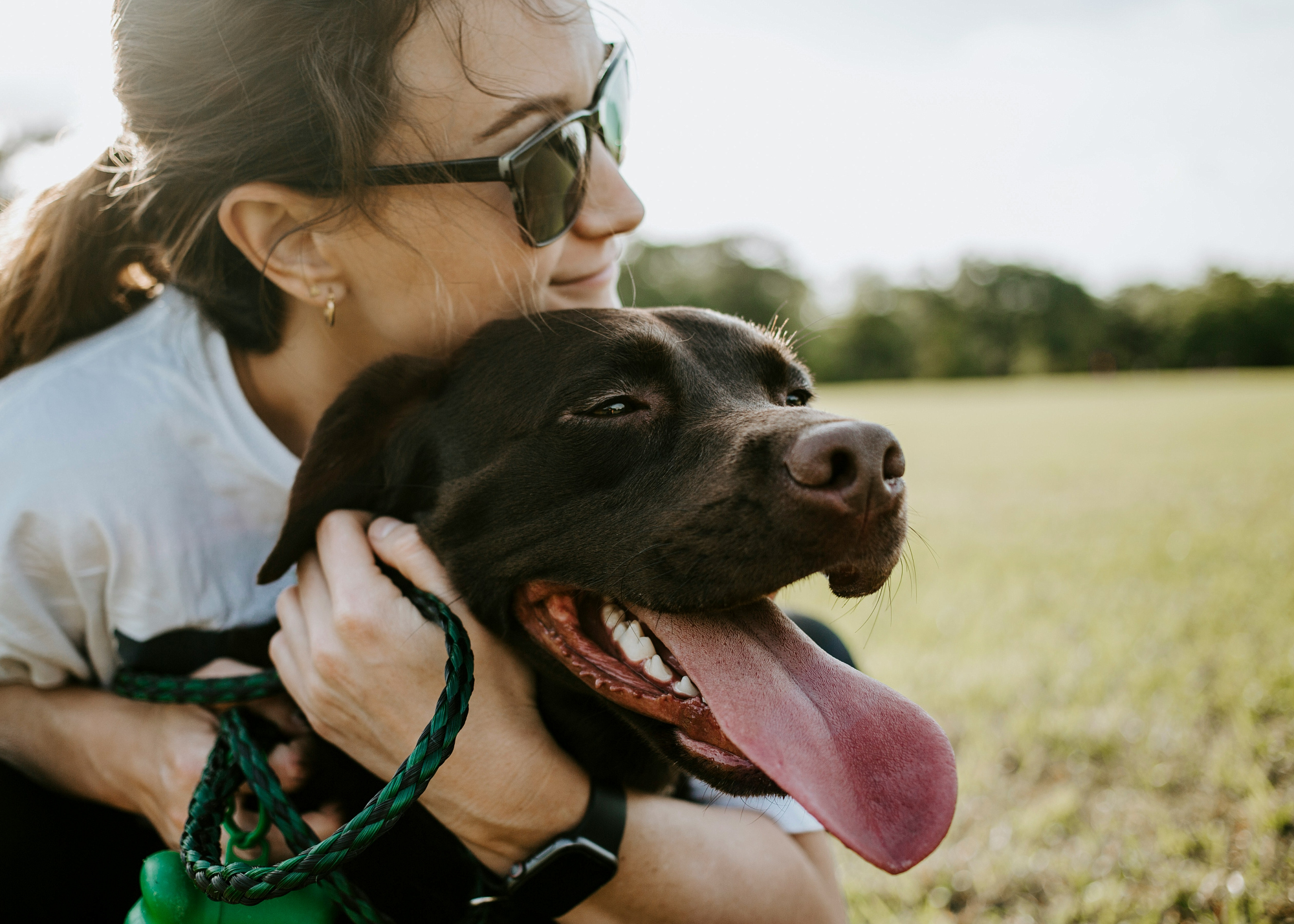 A woman in sunglasses crouches in a field with her arms around a black labrador, which pants happily.